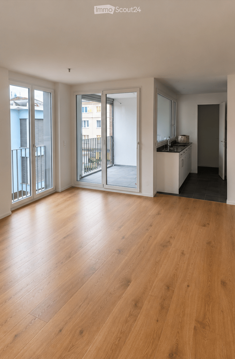 Bright, unfurnished room with wooden flooring, large windows, and sliding glass doors leading to a balcony.