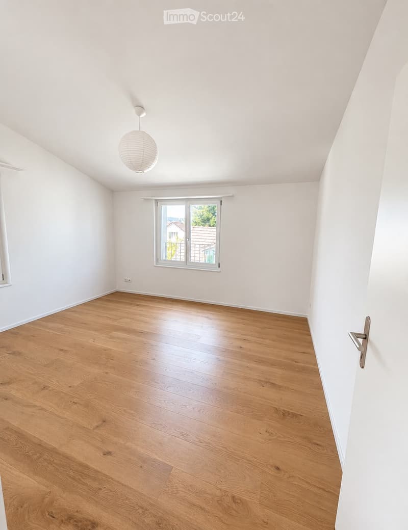 Empty room with light wooden flooring, white walls, and a single window with a view of a neighboring building. A hanging light fixture is visible on the ceiling.