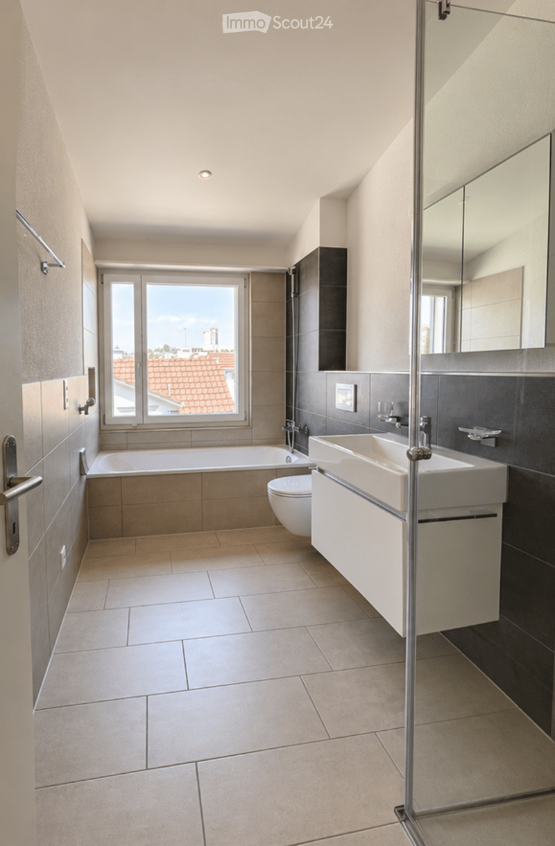 Modern bathroom with a bathtub, toilet, sink, and large mirror. The space features tiled floors and walls, with a window offering a view of the roof.