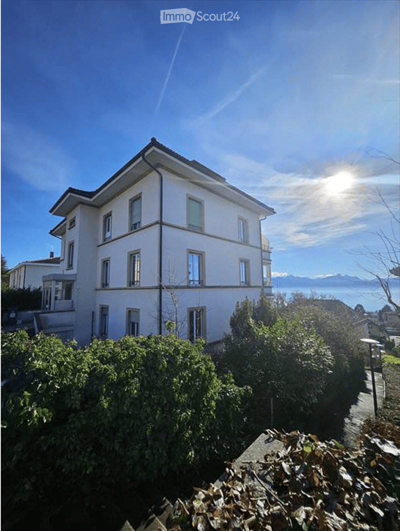A multi-story residential building with white walls, multiple windows, and a view of mountains and a body of water in the background.