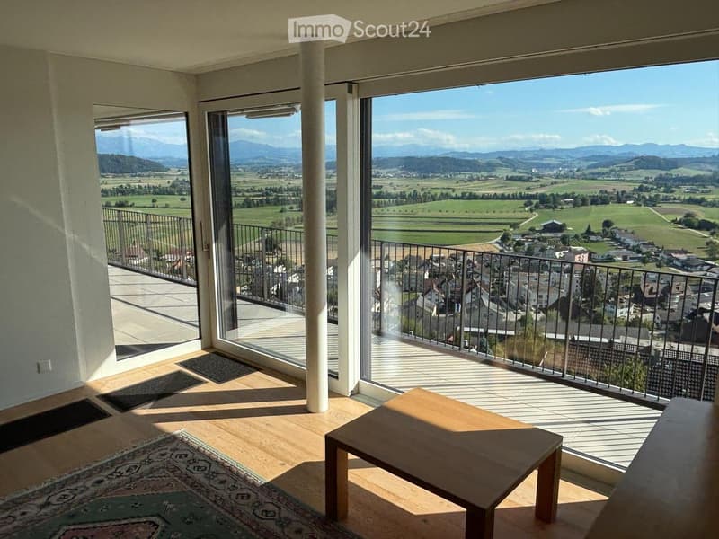 Bright room with wooden flooring, large windows offering scenic views, a small wooden table, and a patterned rug.