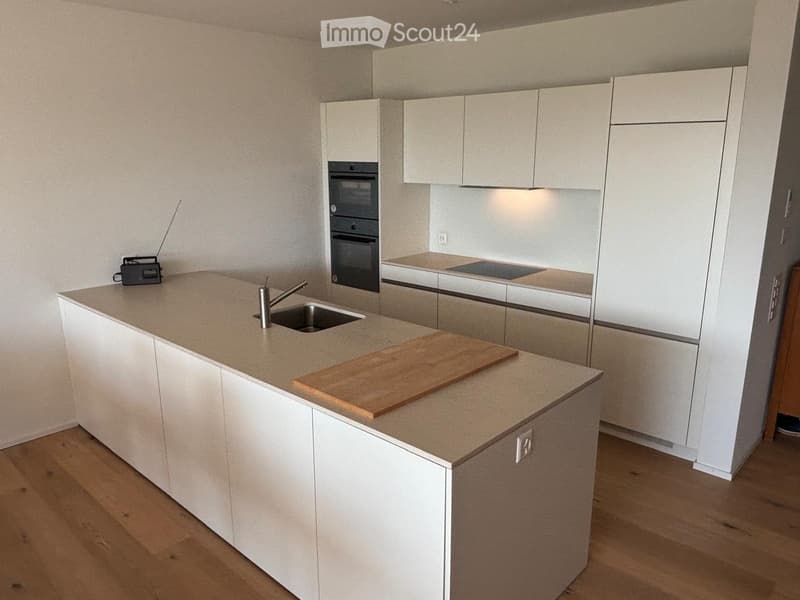 Modern kitchen with white cabinets, light wooden floor, central island with sink and faucet, cutting board, and built-in oven and refrigerator.