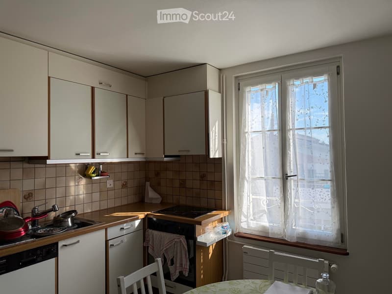 A small kitchen with white cabinets, a tiled backsplash, and a window with sheer curtains. The counter has a sink, stove, and various kitchen items, and there is a small dining area with a table and chairs.