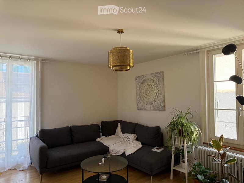 Modern living room with a dark grey sectional sofa, a glass coffee table, a floor lamp, a table lamp, a large painting, and several potted plants near the window.