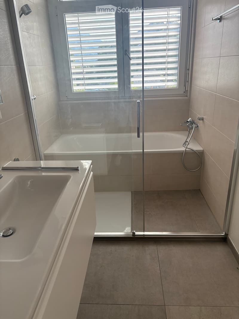 Modern bathroom with a large sink, bathtub, and glass-enclosed shower. The space features light-colored tiles and a window with blinds.