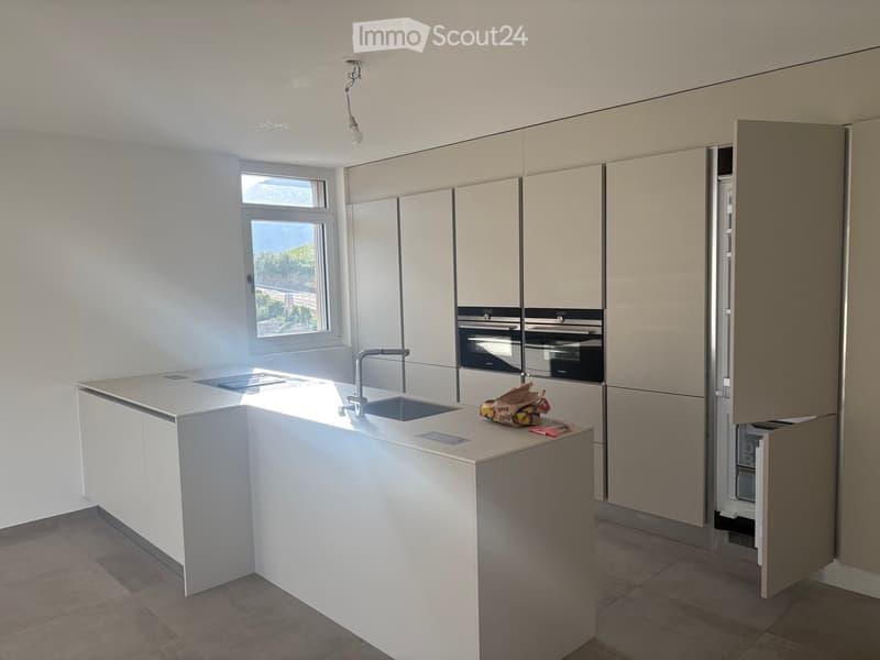 Modern kitchen with white cabinets, a central island, and built-in appliances including an oven and refrigerator. A window provides natural light, and a few grocery bags are placed on the island.