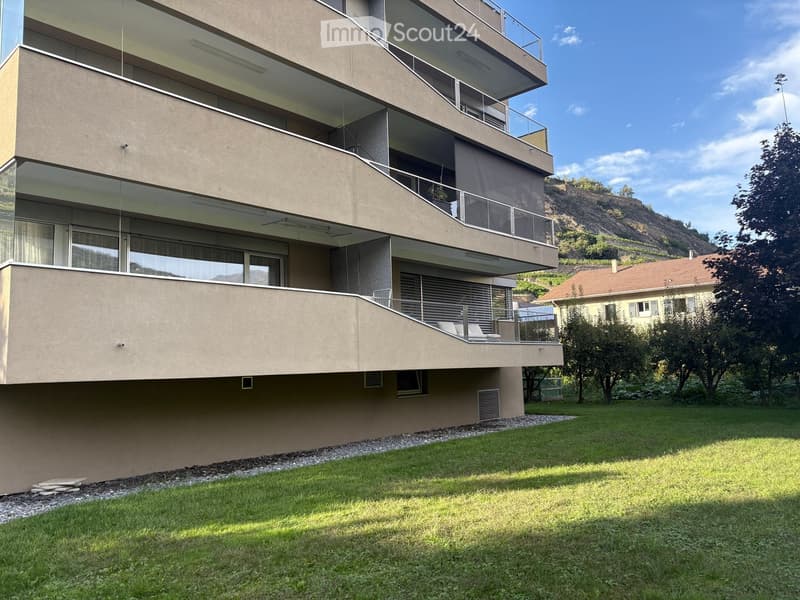 Modern multi-story building with large windows and balconies, surrounded by a well-maintained lawn and trees.