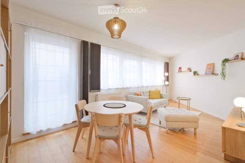 Bright living room with wooden flooring, large windows with white curtains, a sofa with a yellow throw pillow, an ottoman, a dining table with chairs, and a floor lamp.