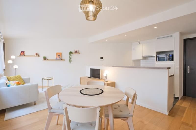 Modern, open-plan living and dining area with a white sofa, small round dining table with four chairs, a television on a stand, and a compact kitchen area with white cabinets and a microwave. Shelves with decorative items and a pendant light are also visi