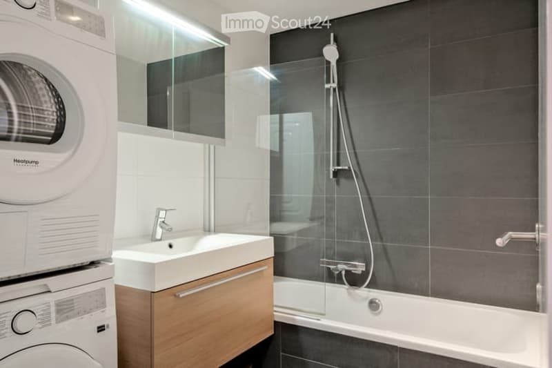 Modern bathroom with a white sink, wooden cabinet, glass-enclosed shower, and a washing machine. The walls are tiled in dark grey, and the floor is also dark grey.