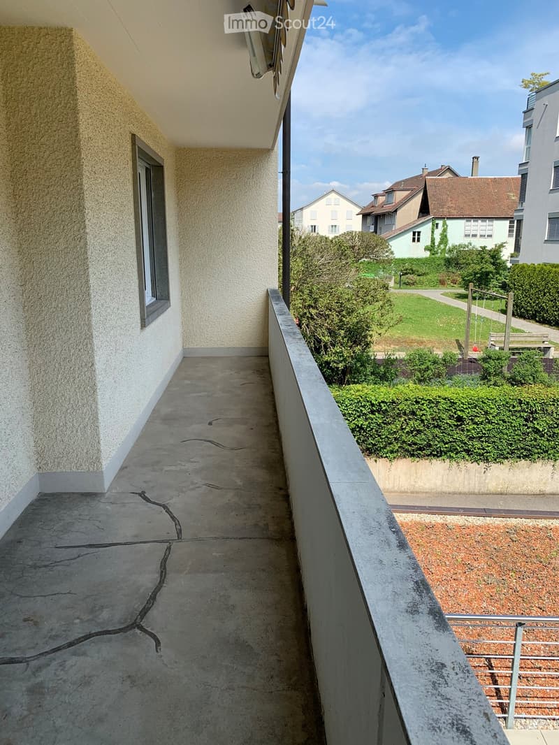 Balcony with concrete floor, white walls, and a metal railing, overlooking a garden and neighboring houses.