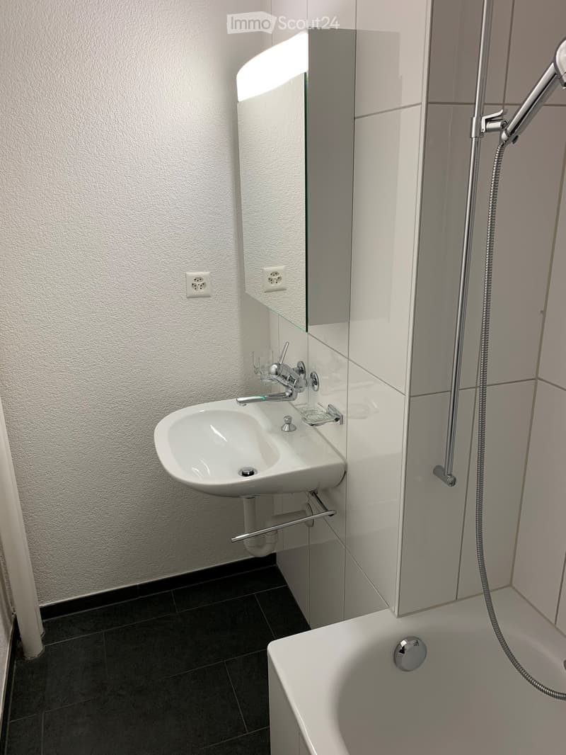 Modern bathroom with a white sink, chrome faucet, bathtub, and glass-enclosed shower area. The walls are white with a textured finish, and the floor is dark tiled.