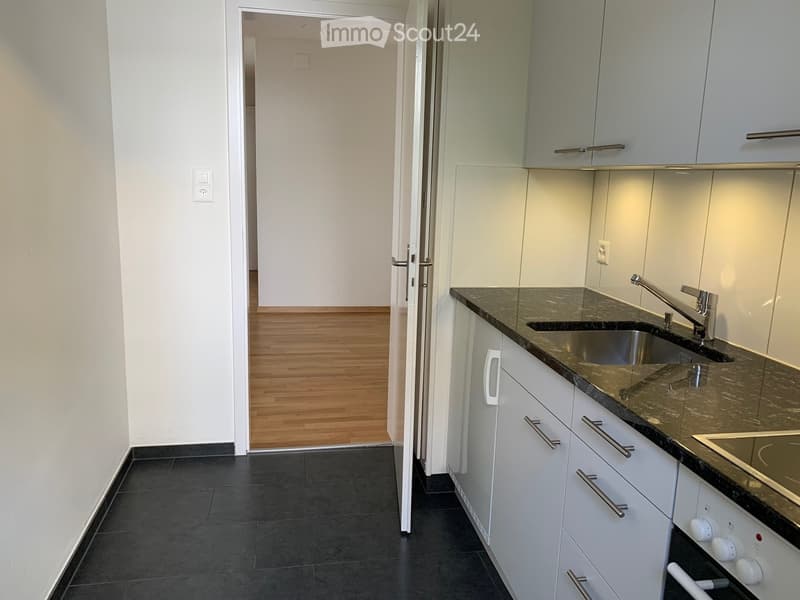 Modern kitchen with white cabinets, black granite countertops, stainless steel sink, and built-in stove. The floor is black tile, and the adjacent room has wooden flooring.