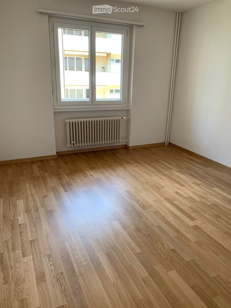 Empty room with wooden flooring, white walls, a radiator, and a large window with a view of neighboring buildings.