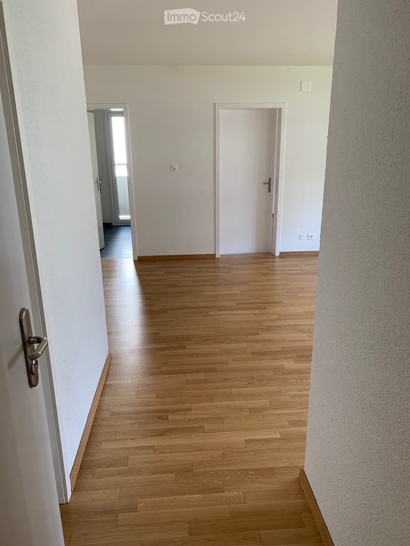 Empty hallway with wooden flooring, white walls, and two doors, one leading to a room and the other to a brightly lit area.