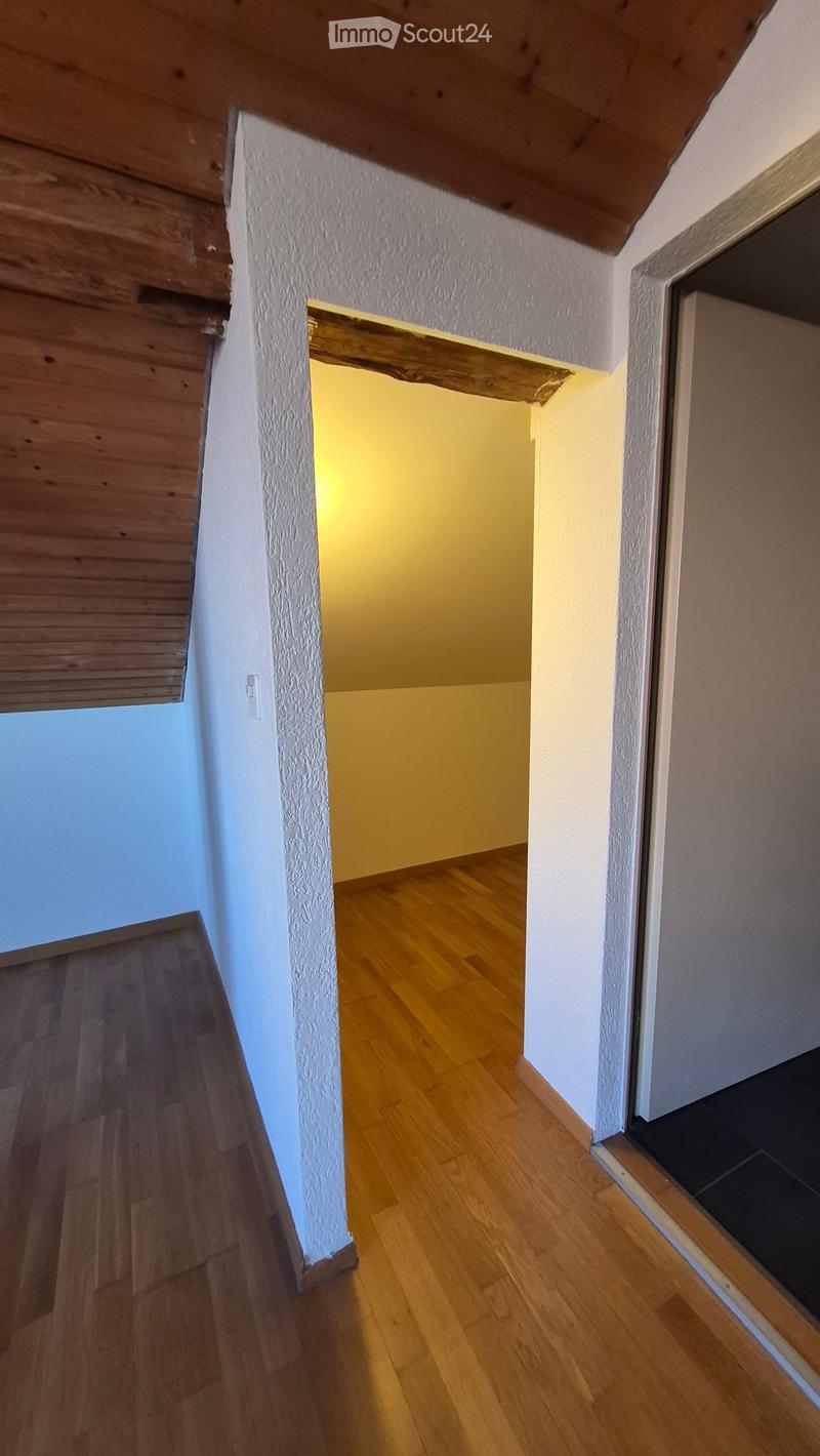 A narrow hallway with wooden flooring and white textured walls, leading to a brightly lit room.