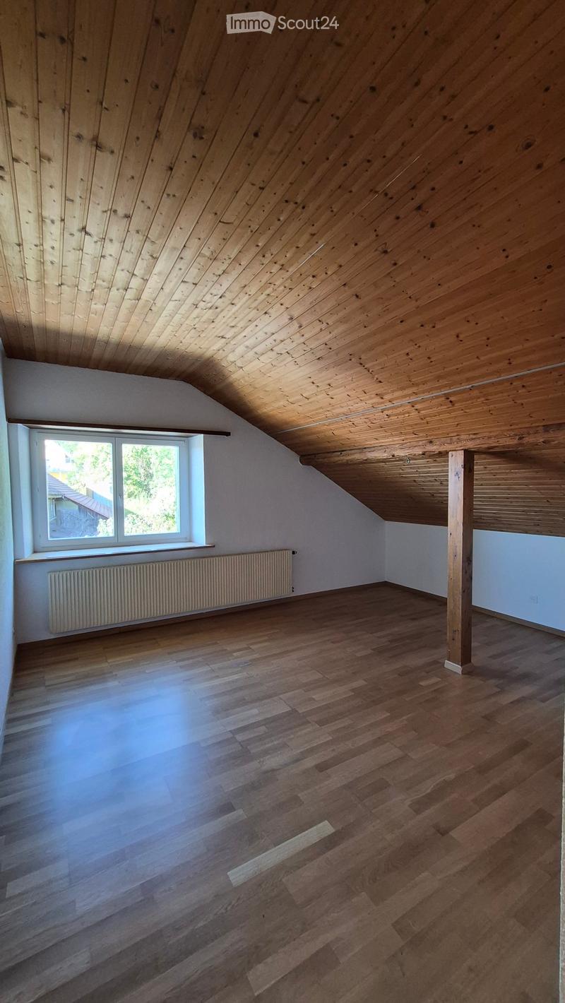 Empty attic room with wooden slatted ceiling, wooden floor, white walls, and a window with a radiator below it.