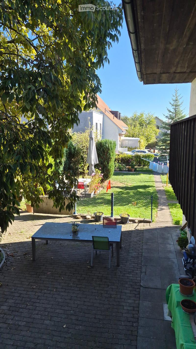 Garden area with a brick-paved patio, a small table with chairs, potted plants, and an umbrella. The garden is enclosed by a fence and features a grassy lawn and trees.