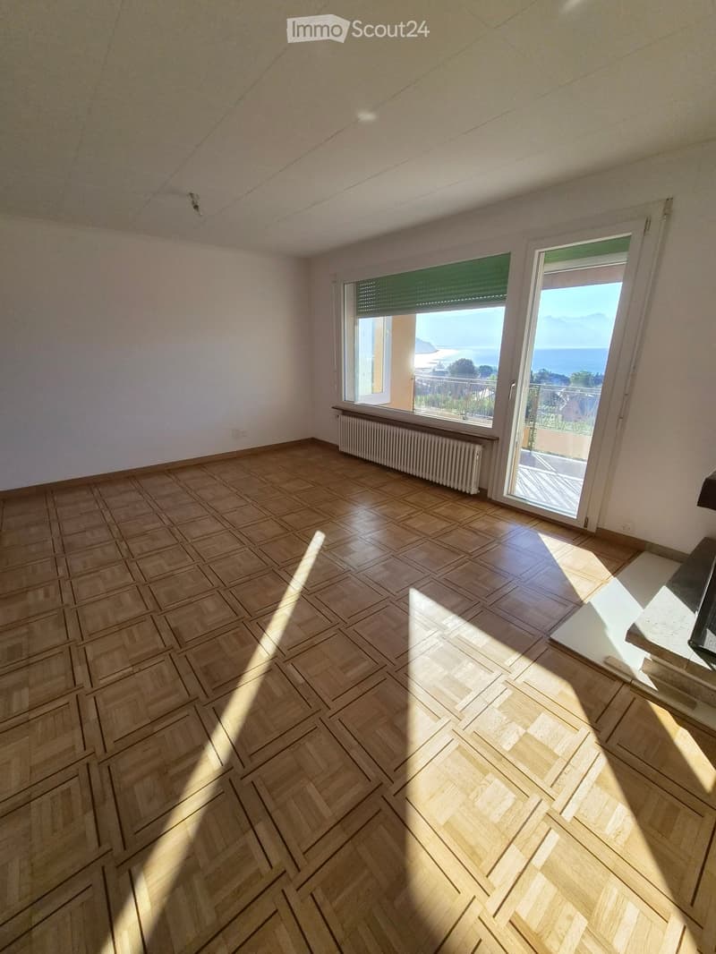 Empty room with wooden herringbone flooring, white walls, large window with blinds, and a radiator below it. The room has a view of the sea through the window.