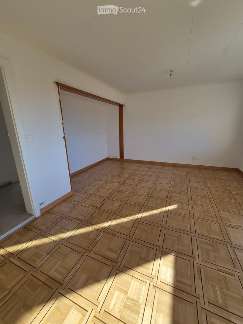 Empty room with light wooden herringbone flooring and white walls, featuring a doorway on the left side.