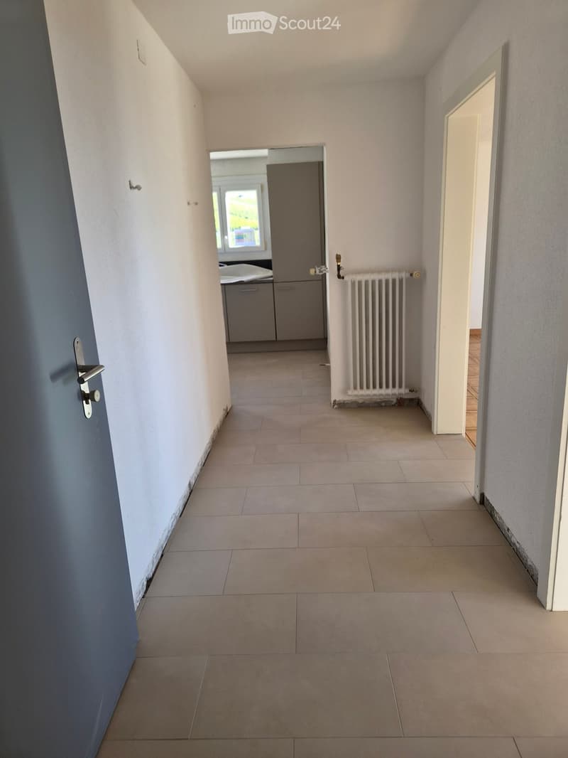 Empty hallway with tiled floor, white walls, and a radiator on the right side. A door leads to a room with a window and cabinets visible in the background.