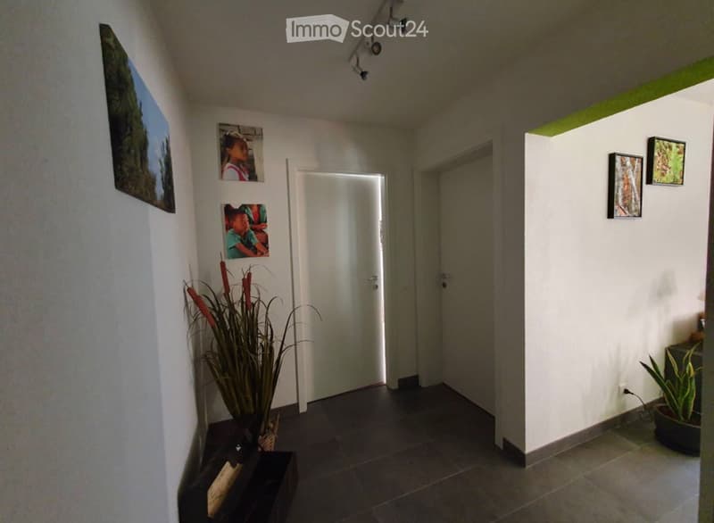 A modern hallway with white walls, dark tiled floor, and several framed pictures. There are two doors, one leading to another room, and small potted plants on either side.