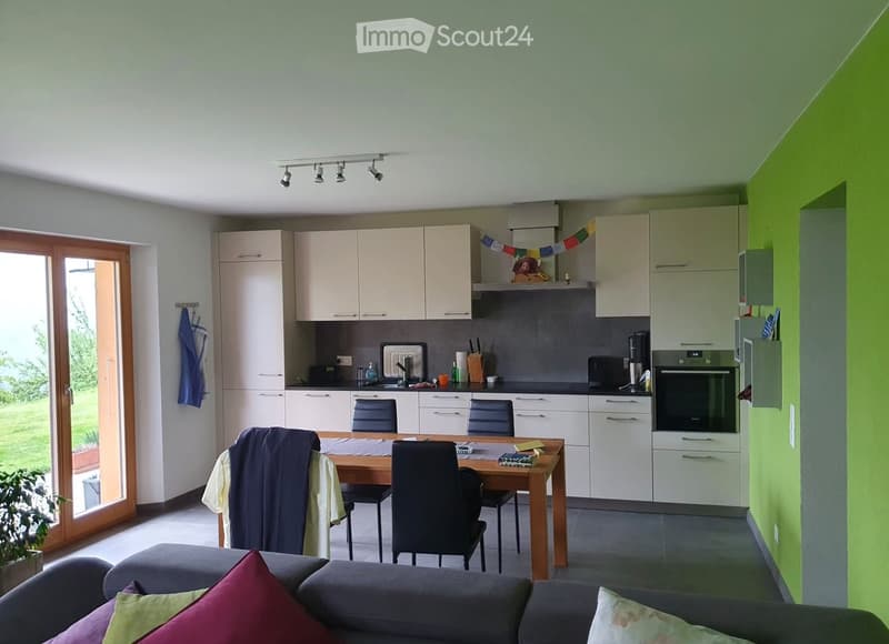 Open-plan living and dining area with a grey sofa, wooden dining table, chairs, and modern kitchen cabinets. The kitchen features an oven, refrigerator, and various appliances.