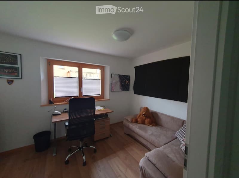 A small, well-lit room with a sofa, a desk, and a chair. The room features a large window with blinds, a screen on the wall, and a plush toy on the sofa.