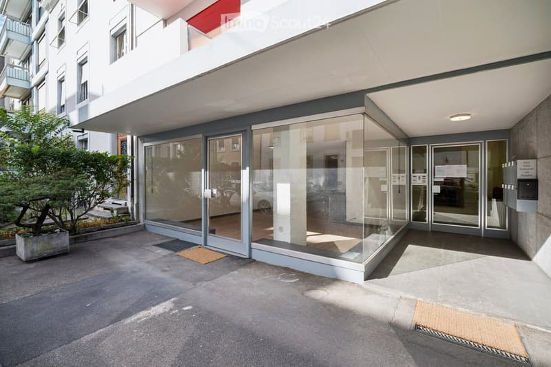 Modern building entrance with large glass doors, a small tree in a planter, and a paved area.