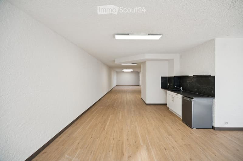 A modern, minimalist kitchen area with light wooden flooring, white walls, and sleek black countertops. The space includes a refrigerator, sink, and cabinets.