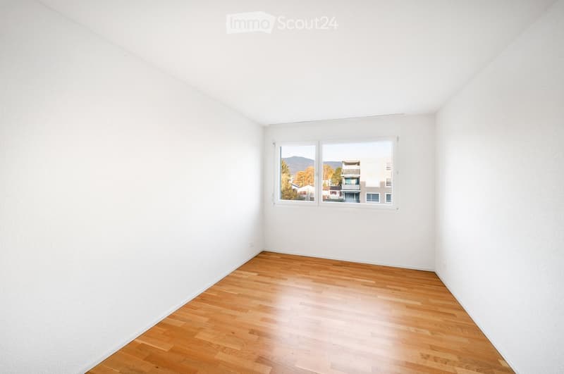 Empty room with white walls and wooden flooring, featuring a large window with a view of buildings and trees.