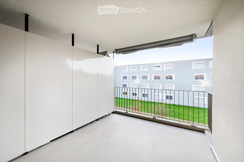 Empty balcony with white walls, metal railing, and a view of a modern apartment building and green lawn.