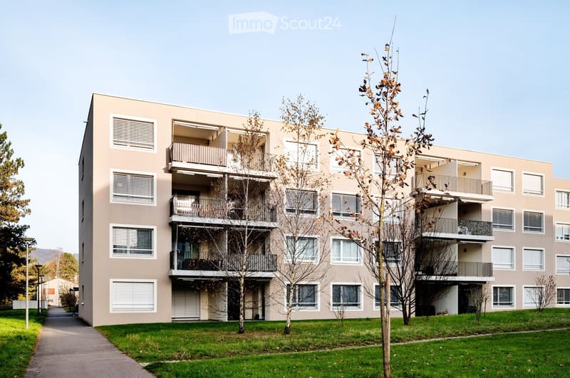 Modern multi-story residential building with beige facade, multiple balconies, and surrounding greenery.