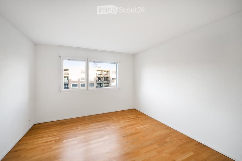 Empty room with white walls, wooden floor, and large window showing a view of neighboring buildings.