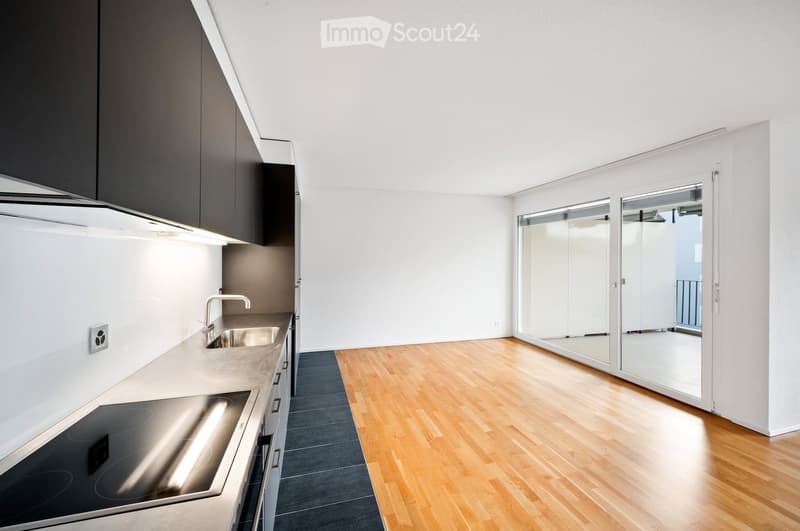 Modern, minimalist kitchen with sleek white cabinets, stainless steel appliances, and a large window leading to a balcony. The space features a wooden floor and a dark grey rug.