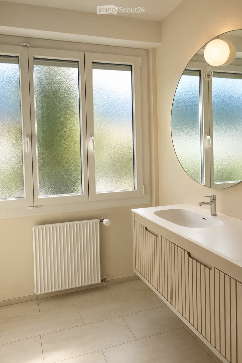 Modern bathroom with a white sink, chrome faucet, large round mirror, and a white radiator under the windows.