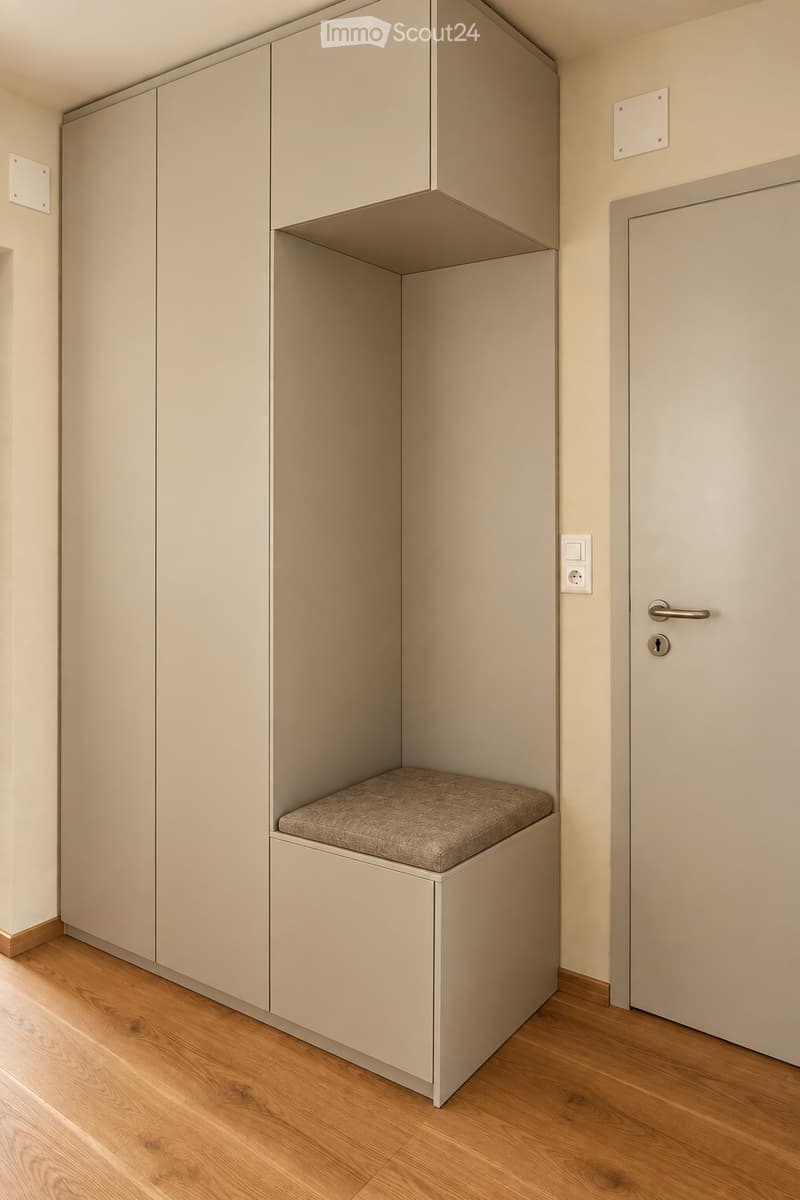 An unfurnished room with light-colored walls, wooden flooring, and built-in storage units with a cushioned seat. A closed door is visible on the right side.