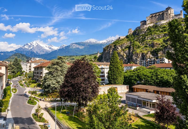 A scenic town view with a castle perched on a hill, surrounded by modern buildings, lush greenery, and snow-capped mountains in the background.