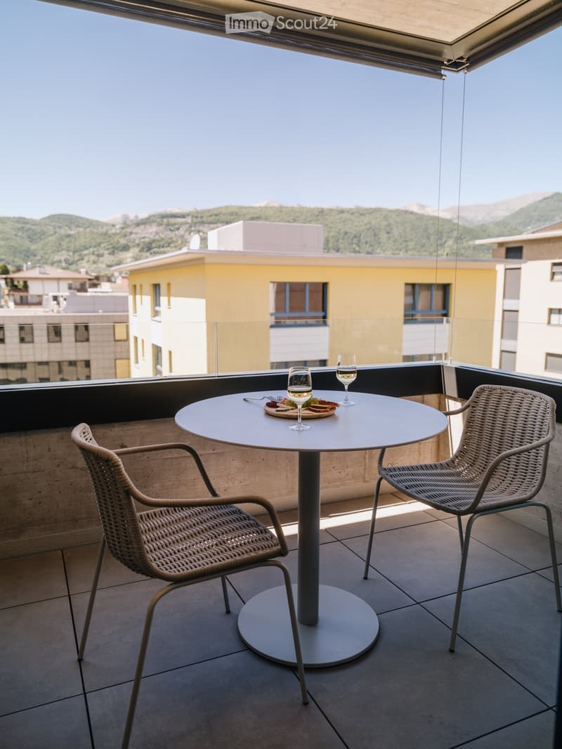 Balcony with a round table, two wicker chairs, and a glass of white wine, overlooking a cityscape with buildings and mountains.