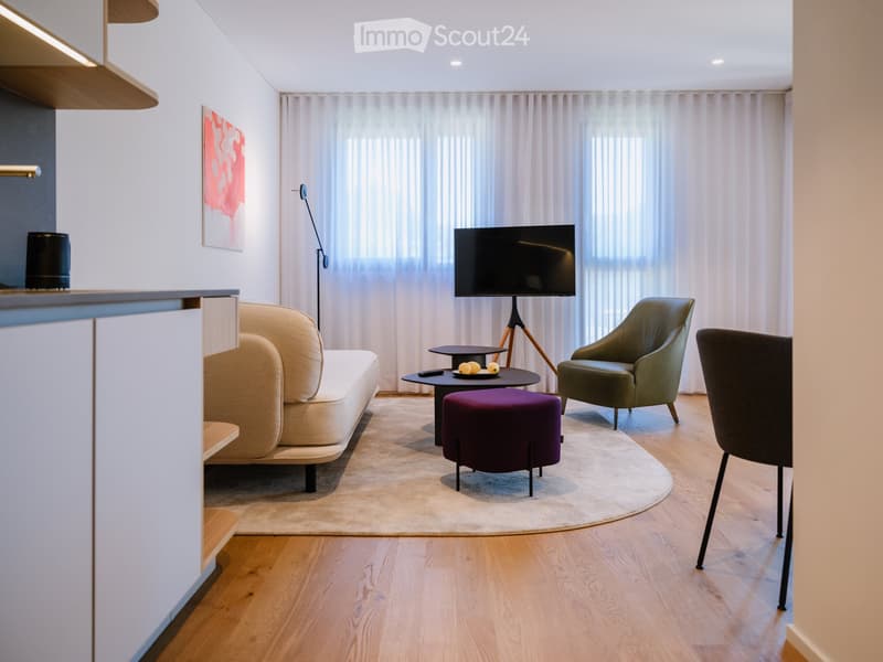 Modern living room with a beige sofa, green armchair, purple ottoman, coffee table, TV on a stand, floor lamp, and large windows with white curtains.