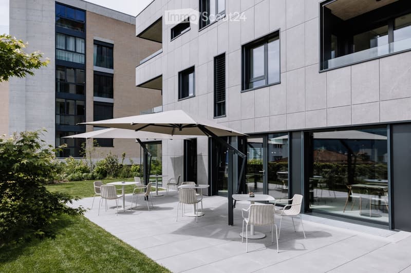Modern outdoor seating area with white tables and chairs, large umbrella, modern building with large windows in the background.
