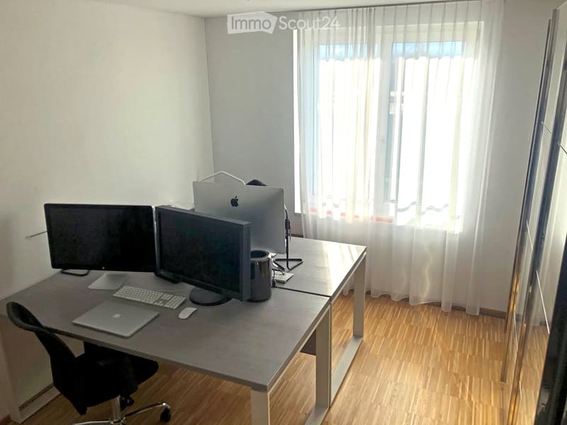 A modern, well-lit room with a large wooden desk, multiple monitors, a laptop, a chair, and a cup. The room has large windows with sheer curtains and wooden flooring.