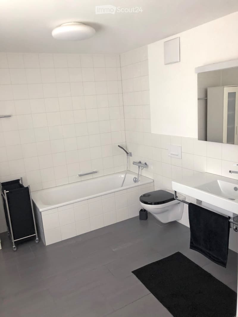 Modern bathroom with white tiled walls, a bathtub, toilet, sink, and a mirror. Includes a black towel on the sink and a small black rug on the floor.