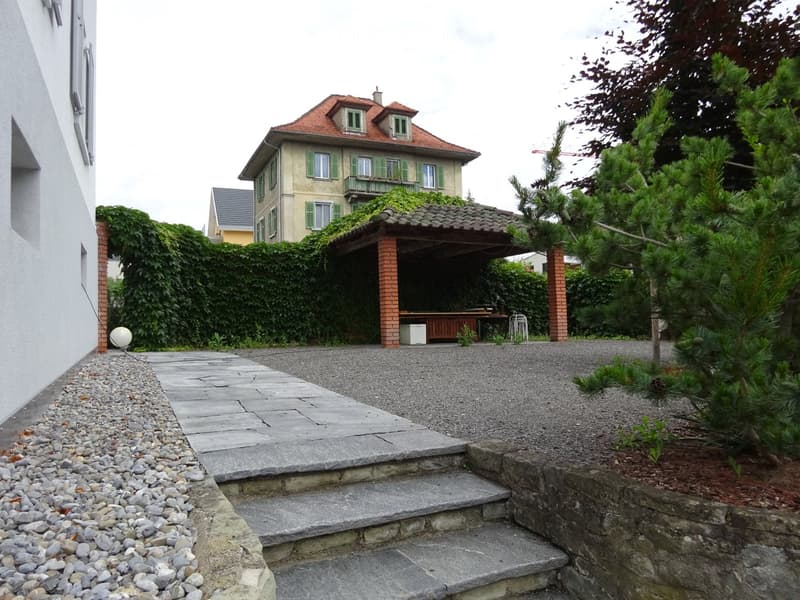 Garden area with gravel pathways, stone steps, a covered outdoor seating area, and surrounding greenery including trees and shrubs.