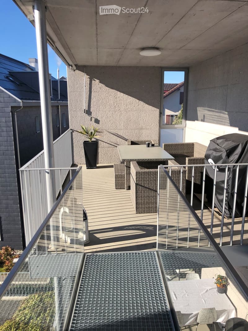 Balcony with a table, chairs, and a potted plant, featuring a modern glass railing and a view of the building exterior.