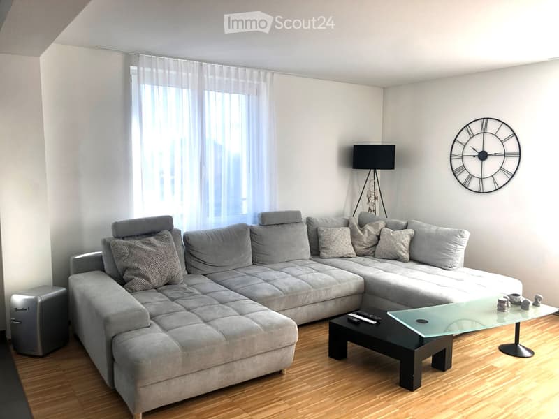 Modern living room with a grey sectional sofa, glass coffee table, floor lamp, and a large wall clock. The room features wooden flooring and large windows with sheer curtains.