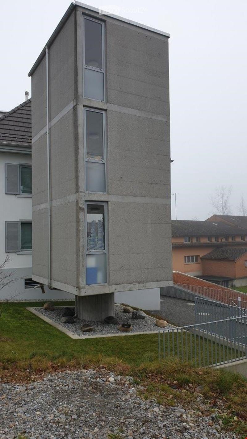Modern, raised concrete building with large windows and a minimalist design, elevated on stilts with a gravel base.