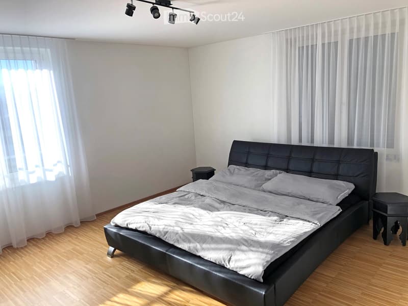 Modern bedroom with a large black leather bed, grey bedding, two nightstands, and sheer white curtains on large windows.