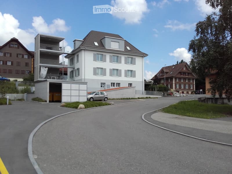 Modern two-story building with white facade, grey roof, and balcony on the upper level. The entrance features a covered parking area with a car parked inside. The surrounding area includes a curved driveway, small landscaped garden, and neighboring houses
