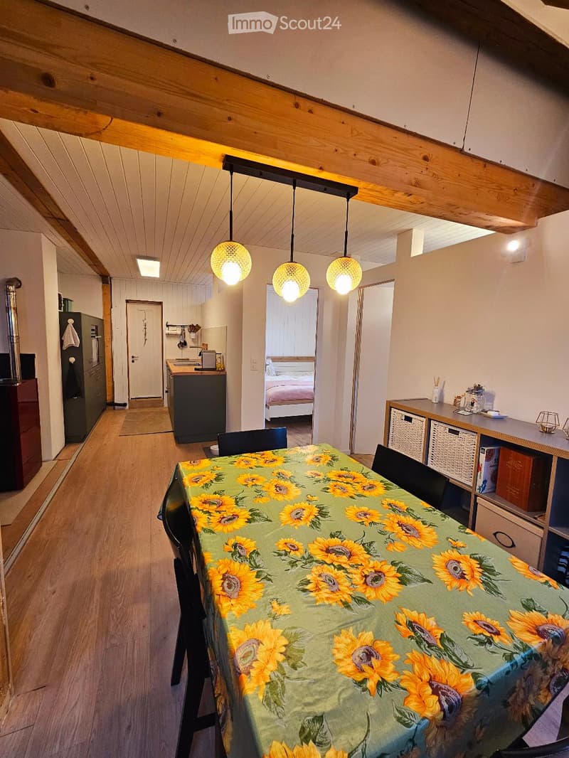 A cozy, well-lit room with wooden floors and exposed wooden beams. Features a dining table with a sunflower-patterned tablecloth, surrounded by chairs. A kitchen area is visible with a refrigerator, counter, and cabinets. A bedroom area with a bed is also